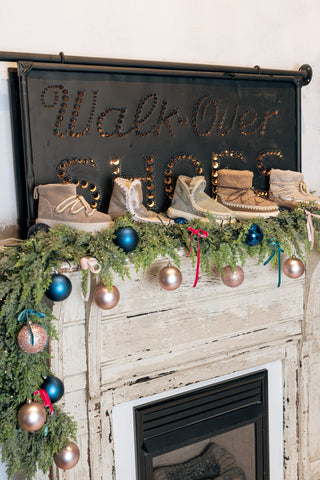 A line of fuzzy boots sit over a fireplace with a green garland and ornaments
