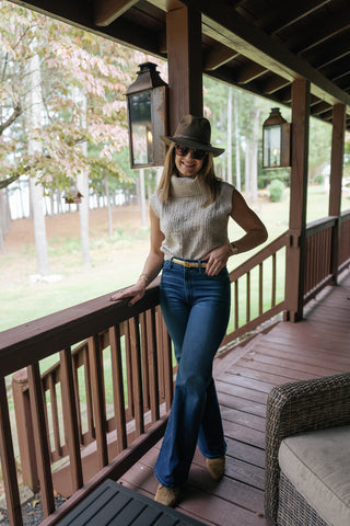 Caleigh poses on the wooden deck of a lake house. She is wearing a brown wide-brim hat, short-sleeved turtleneck sweater, and boot-cut dark jeans. Weathered lanterns and trees slope down to the lake behind her.