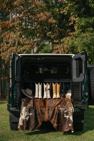 Three pairs of knee-high cowboy boots and a wide-brimmed hat lay on top of a cowhide rug, draped through the open doors of a large SUV's trunk