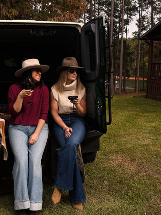 Caleigh and Nikki wear fall clothes, sip wine, and sit in the back of a large SUV by a lakeside