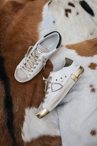 Two white, gold, and pewter colored Philippe Model sneakers lay on top of a brown, black, and white cowhide rug
