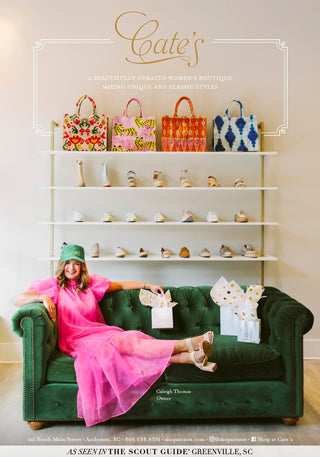 Caleigh Thomas, owner of Cate's, is wearing a pink dress and Cate's trucker hat while lounging on a green velvet couch in front of shelves of shoes and bags. 